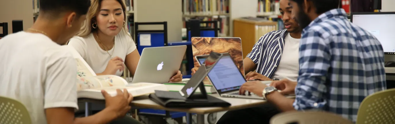 Four students collaborating over laptops and books at a library table