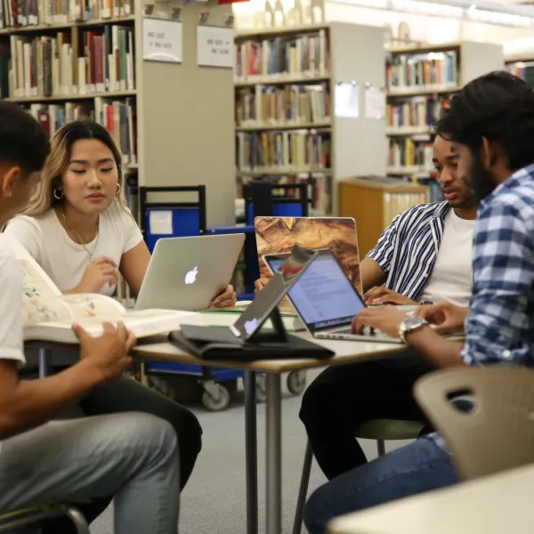 Four students working on laptops at a table in front of library stacks
